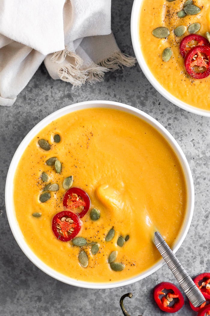 Overhead shot of creamy pumpkin soup garnished with pumpkin seeds, chili pepper, and pepper. Next to it is more sliced peppers, another bowl, and a white linen.