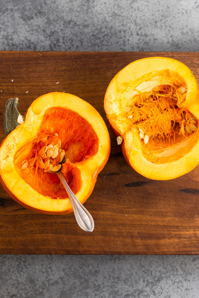 A small sugar pumpkin cut in half sitting on a cutting board. The is a spoon in one of the halves.