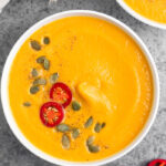 Overhead shot of Instant Pot pumpkin soup garnished with pumpkin seeds, chili pepper, and pepper. Next to it is more sliced peppers and another bowl.