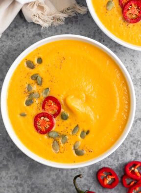 Overhead shot of Instant Pot pumpkin soup garnished with pumpkin seeds, chili pepper, and pepper. Next to it is more sliced peppers and another bowl.