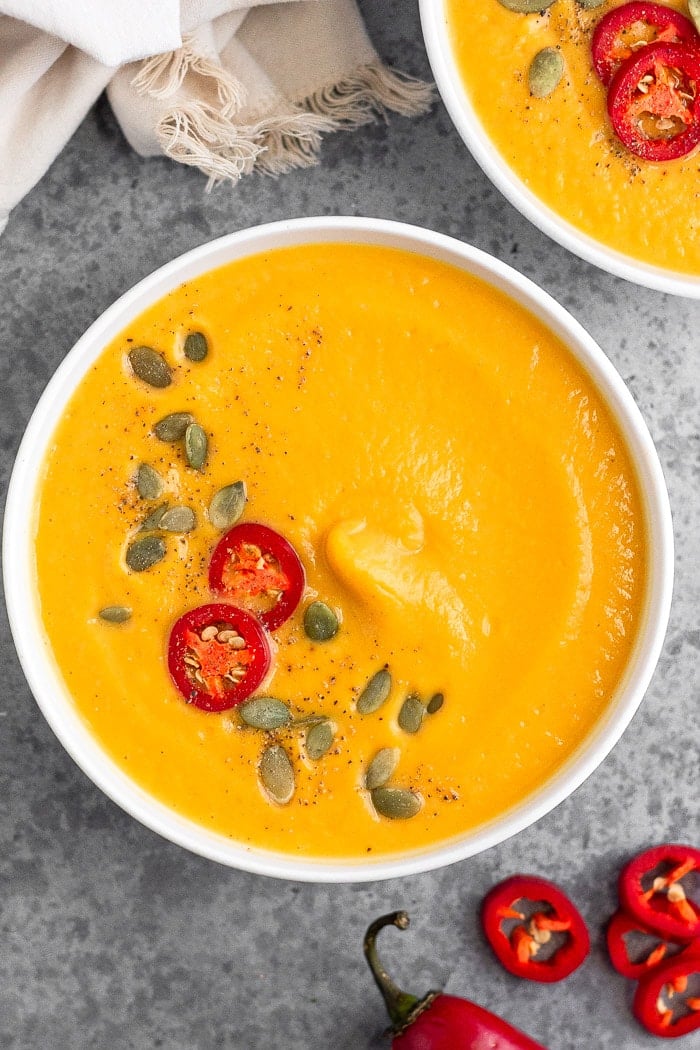 Overhead shot of Instant Pot pumpkin soup garnished with pumpkin seeds, chili pepper, and pepper. Next to it is more sliced peppers and another bowl.