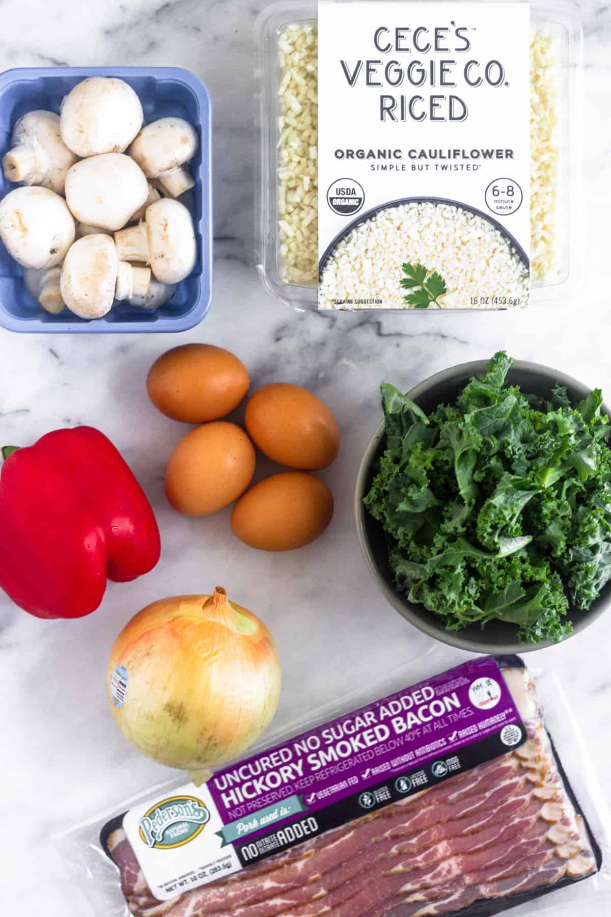 Ingredients to make paleo breakfast fried cauliflower rice on a white counter - mushrooms, cauliflower rice, chopped kale, bacon, onion, red bell pepper, and eggs.