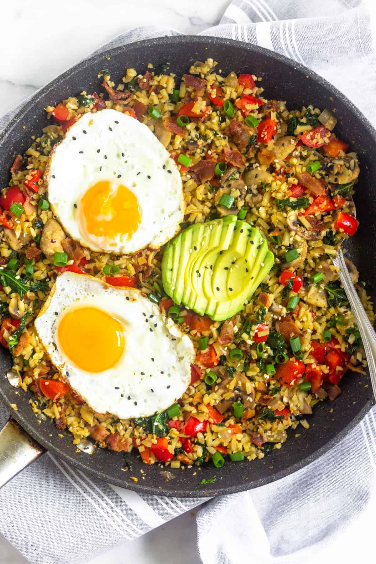 Large saute pan filled with whole30 and paleo breakfast fried rice topped with two fried eggs, sliced avocado, and sesame seeds