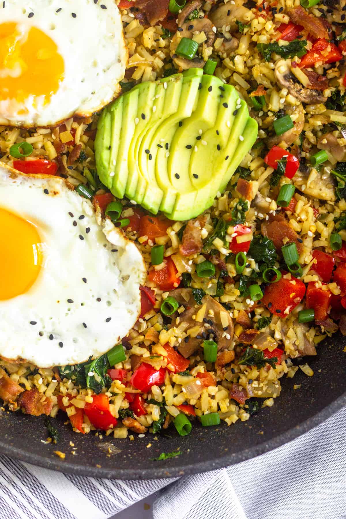Close up of a large black pan filled with paleo breakfast fried rice topped with fried eggs and sliced avocado