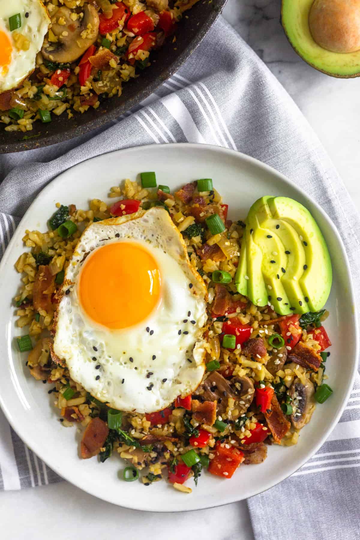 White plate with low carb paleo breakfast fried rice topped with a fried egg and sliced avocado. It is sitting on a towel next to a pan of more fried rice and half and avocado