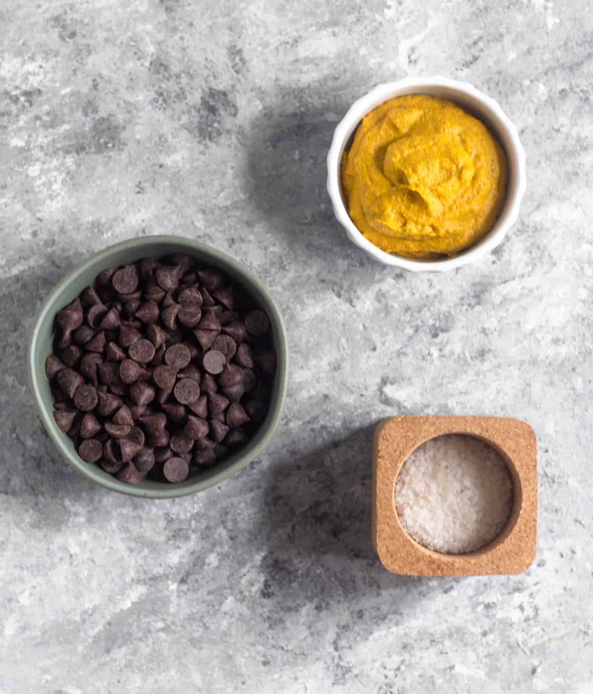 A bowl of chocolate chips, a bowl of pumpkin pie butter, and a container of sea salt on a grey surface