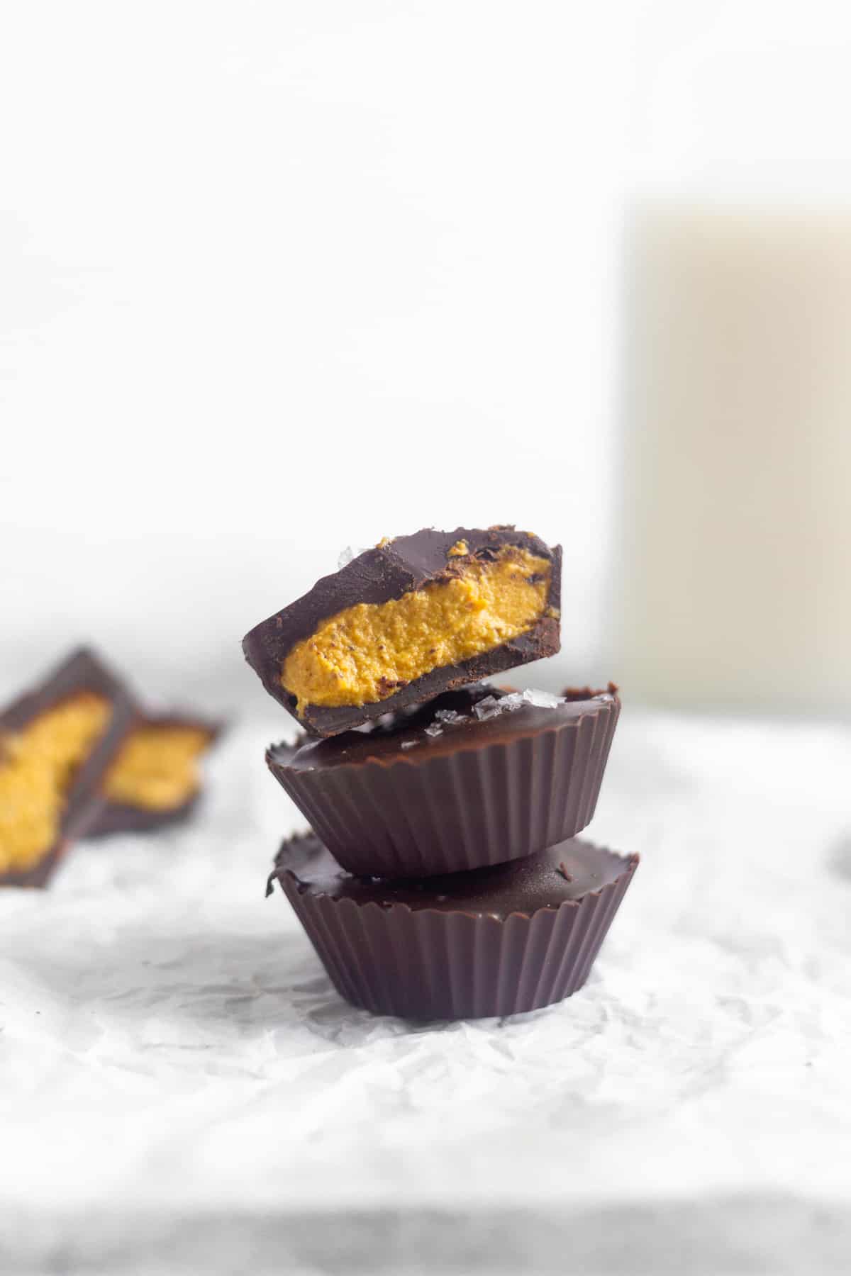 Three pumpkin pie chocolate cups stacked on top of each other. The top one is cut in half. Behind them are more cut open chocolate ups and a jar of milk.