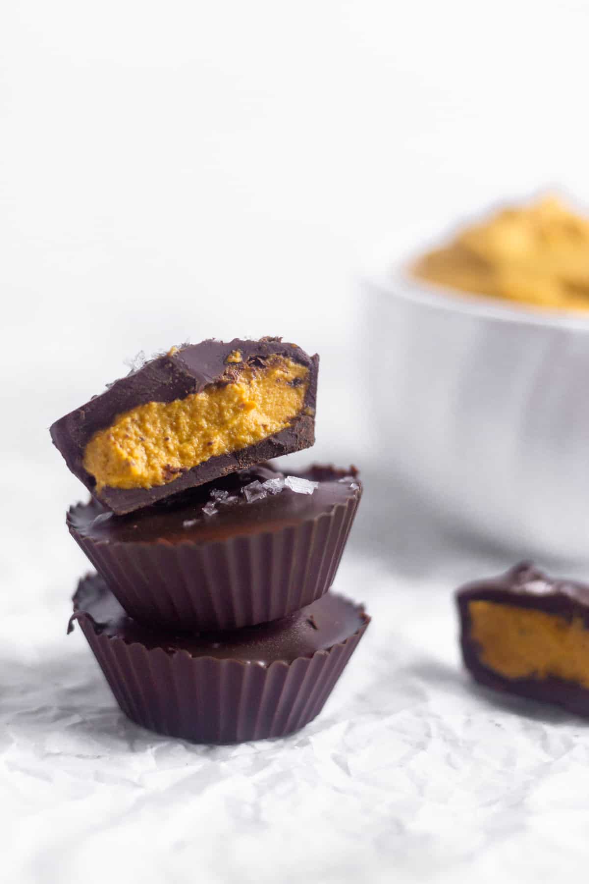 Three pumpkin pie chocolate cups on top of each other. The top one is cut in half with the other half next to the stack. A bowl of pumpkin pie butter is behind them
