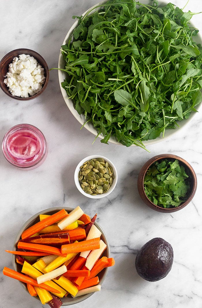 Overhead shot of a bowl filled with arugula, a bowl of cilantro leaves, bowl of cut up rainbow carrots, bowl of pumpkin seeds, jar of pickled onions, and a bowl of feta.