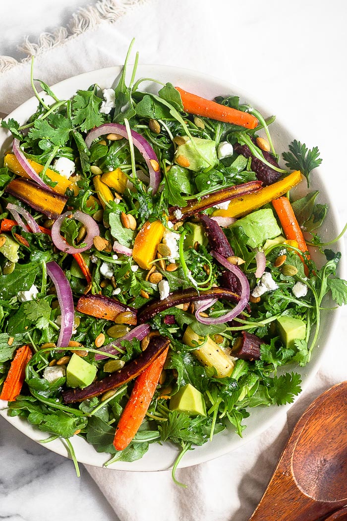 Large white bowl of carrot salad with arugula, feta, avocado, and onions. Next to it is two wooden tongs.