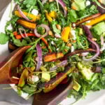 Overhead shot of a bowl of roasted carrot salad with arugula, feta, avocado, and onions. Two wooden tongs are reaching in to take some.