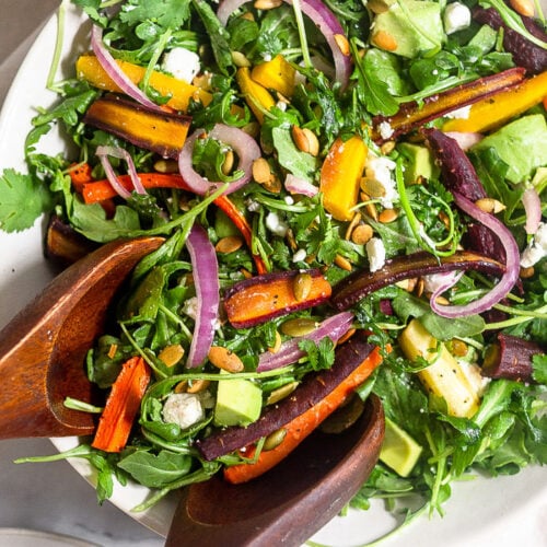 Overhead shot of a bowl of roasted carrot salad with arugula, feta, avocado, and onions. Two wooden tongs are reaching in to take some.