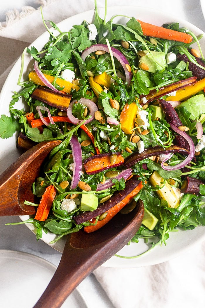 Overhead shot of a bowl of roasted carrot salad with arugula, feta, avocado, and onions. Two wooden tongs are reaching in to take some.