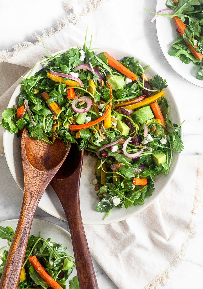 Salad with roasted carrots, arugula, onion, feta, and avocado in a large white bowl. Some is missing from the bowl and two tongs are in there. Next to it is two plates filled with the salad.