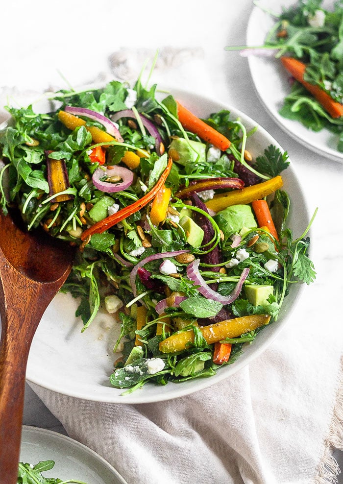 Roasted carrot and arugula salad in a large bowl with some taken out and on a plate in the background. There is also a wooden tong in the salad bowl.