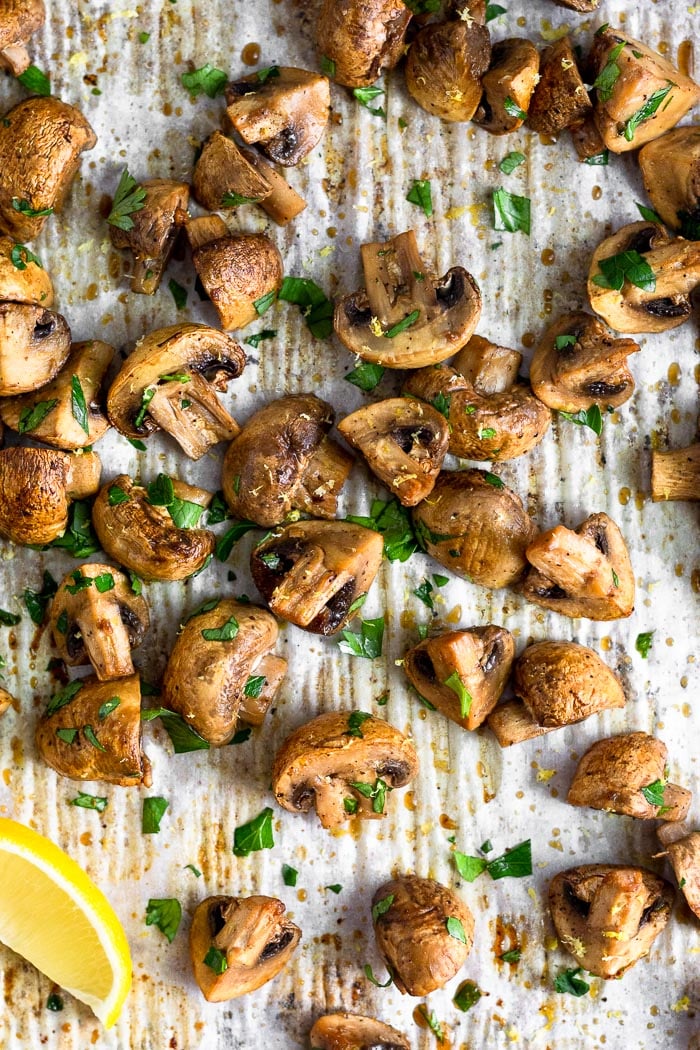 Overhead shot of a baking sheet filled with oven roasted mushrooms topped with fresh chopped parsley. There is also a lemon wedge on the pan.