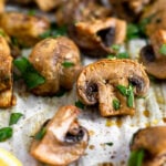 Close up of a balsamic roasted mushrooms on a baking sheet surrounded by more mushrooms. They are topped with fresh parsley.