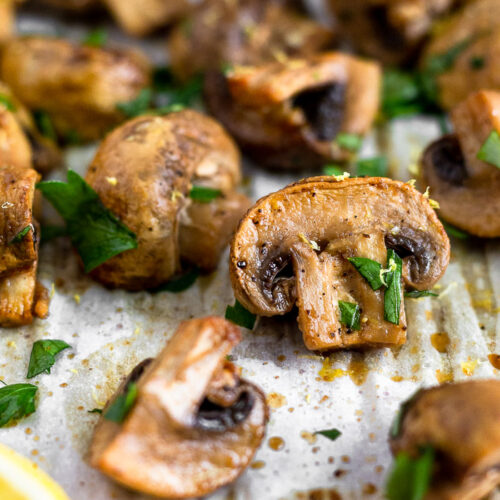 Close up of a balsamic roasted mushrooms on a baking sheet surrounded by more mushrooms. They are topped with fresh parsley.