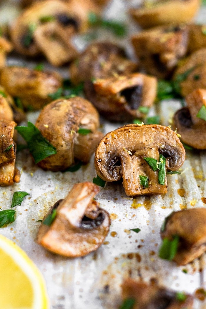 Close up of a balsamic roasted mushrooms on a baking sheet surrounded by more mushrooms. They are topped with fresh parsley.