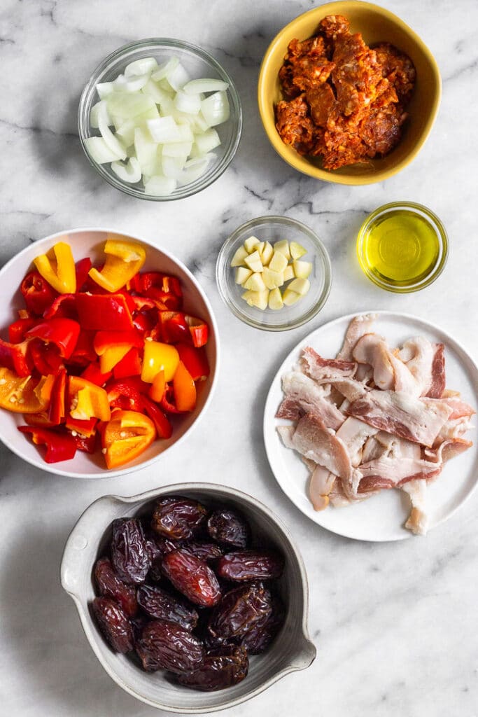 Overhead shot of a small bowl of chopped onion, small bowl of chorizo, small bowl of olive oil, small bowl of chopped garlic, plate of pieces of bacon, bowl of medjool dates, and a bowl of chopped red, yellow, and orange peppers.