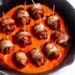 Small cast iron skillet bacon-wrapped chorizo-stuffed dates sitting on top of red pepper sauce.