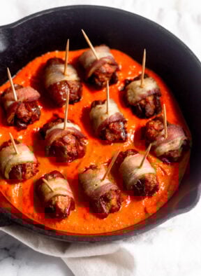 Small cast iron skillet bacon-wrapped chorizo-stuffed dates sitting on top of red pepper sauce.