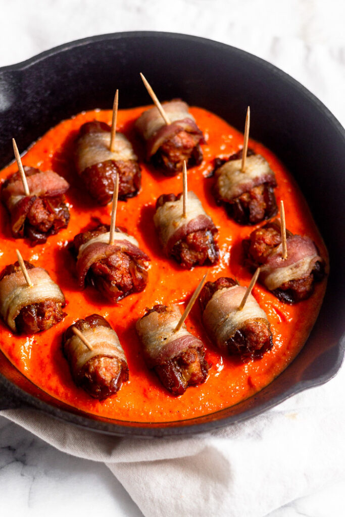 Small cast iron skillet bacon-wrapped chorizo-stuffed dates sitting on top of red pepper sauce.