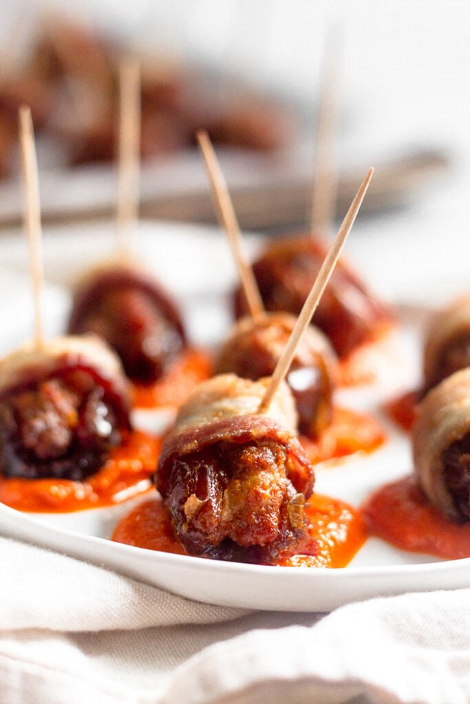 Plate with small dollops of red pepper sauce. On top of the sauce are bacon wrapped stuffed dates. In the background is a sheet pan of more bacon wrapped dates.
