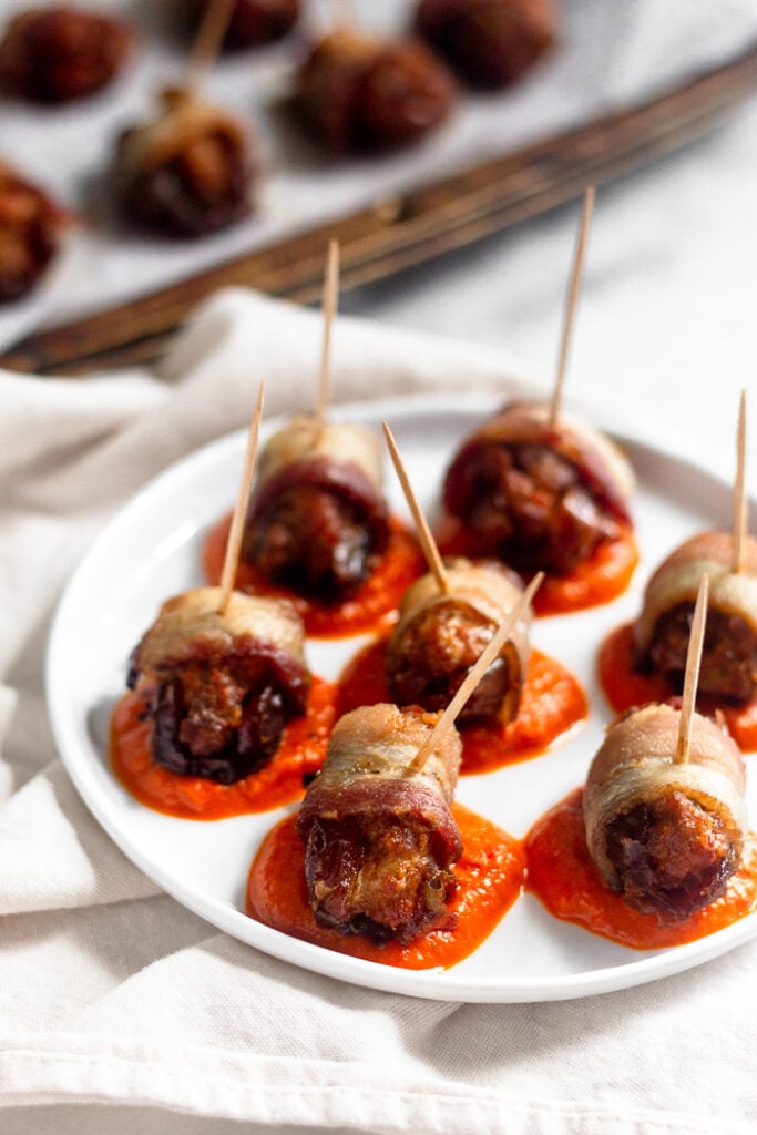 Small white plate with dollops of red pepper sauce on it. On top of the sauce are chorizo-stuffed bacon-wrapped dates. In the background is a sheet pan of more bacon wrapped dates.