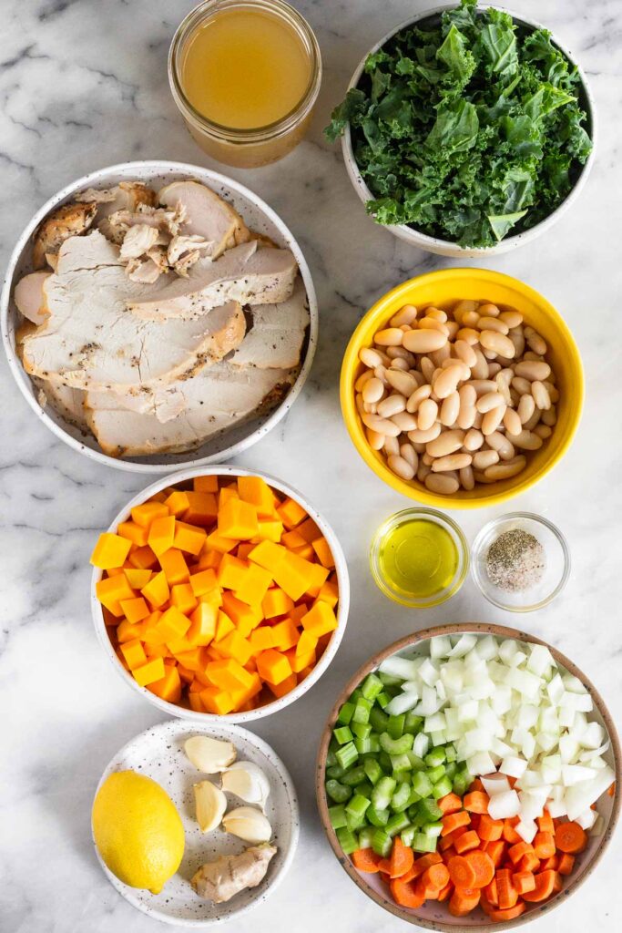 White marble counter with a jar of broth, a bowl of kale, a bowl of white beans, a small bowl of olive oil, a small bowl of salt and pepper, a bowl of diced celery, onions, and carrots, a small plate with a whole lemon, four garlic cloves, and a knob of fresh ginger, a bowl of diced butternut squash, and a bowl of sliced roasted turkey.