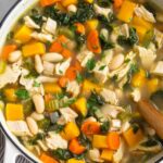 A large pot of leftover turkey soup with butternut squash, kale, veggies, and beans. It is topped with fresh herbs and there is a wooden spoon in the pot.