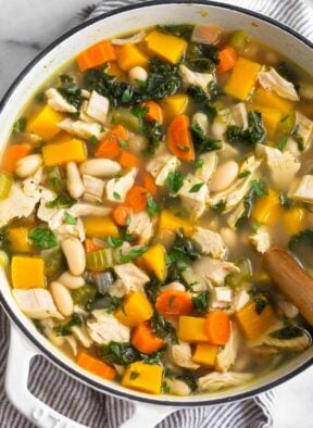 A large pot of leftover turkey soup with butternut squash, kale, veggies, and beans. It is topped with fresh herbs and there is a wooden spoon in the pot.