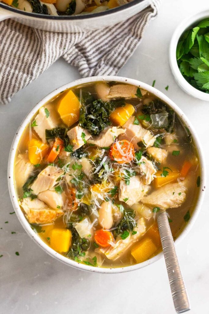A bowl of leftover turkey soup with veggies and beans topped with fresh herbs and parmesan cheese. A spoon is sticking out of the bowl. Around the bowl is a ramekin of fresh herbs, a striped kitchen towel, and a pot of more soup.