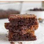 A stack of 3 gingerbread blondies on a piece of parchment paper. Behind them is a baking dish of more blondies, a bowl of chocolate chips, and a jar of vanilla.