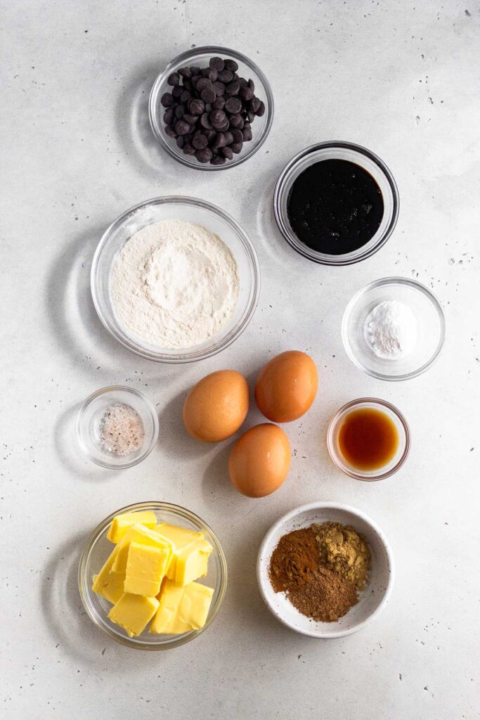 White counter top with a bowl of chocolate chips, a bowl of molasses, a bowl of baking powder, a bowl of vanilla extract, a bowl of spices, a bowl of butter, 3 eggs, a bowl of salt, and a bowl of flour.