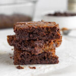 A stack of 3 gingerbread blondies on a piece of parchment paper. Behind them is a baking dish of more blondies, a bowl of chocolate chips, and a jar of vanilla.