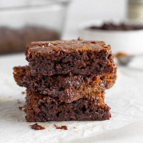 A stack of 3 gingerbread blondies on a piece of parchment paper. Behind them is a baking dish of more blondies, a bowl of chocolate chips, and a jar of vanilla.