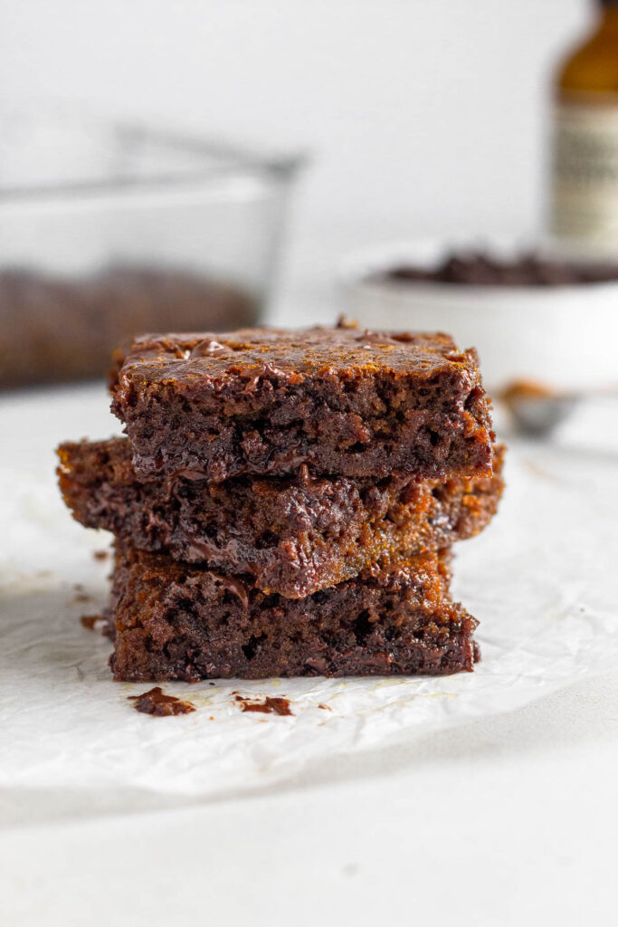 A stack of 3 gingerbread blondies on a piece of parchment paper. Behind them is a baking dish of more blondies, a bowl of chocolate chips, and a jar of vanilla.