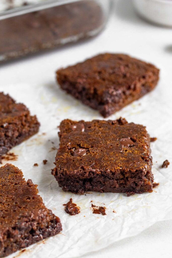Gingerbread blondies with chocolate chips on a piece of parchment paper. There are chocolate crumbs on the parchment paper and behind it is a baking dish with more blondies.