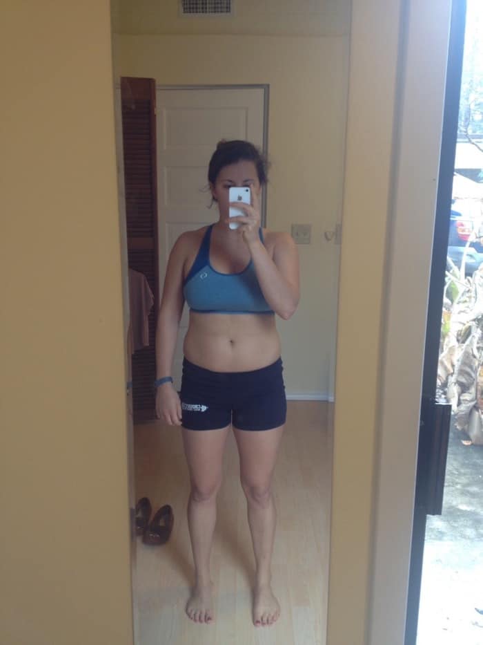 Girl in spandex shorts and a sports bra taking a selfie in front of a mirror.