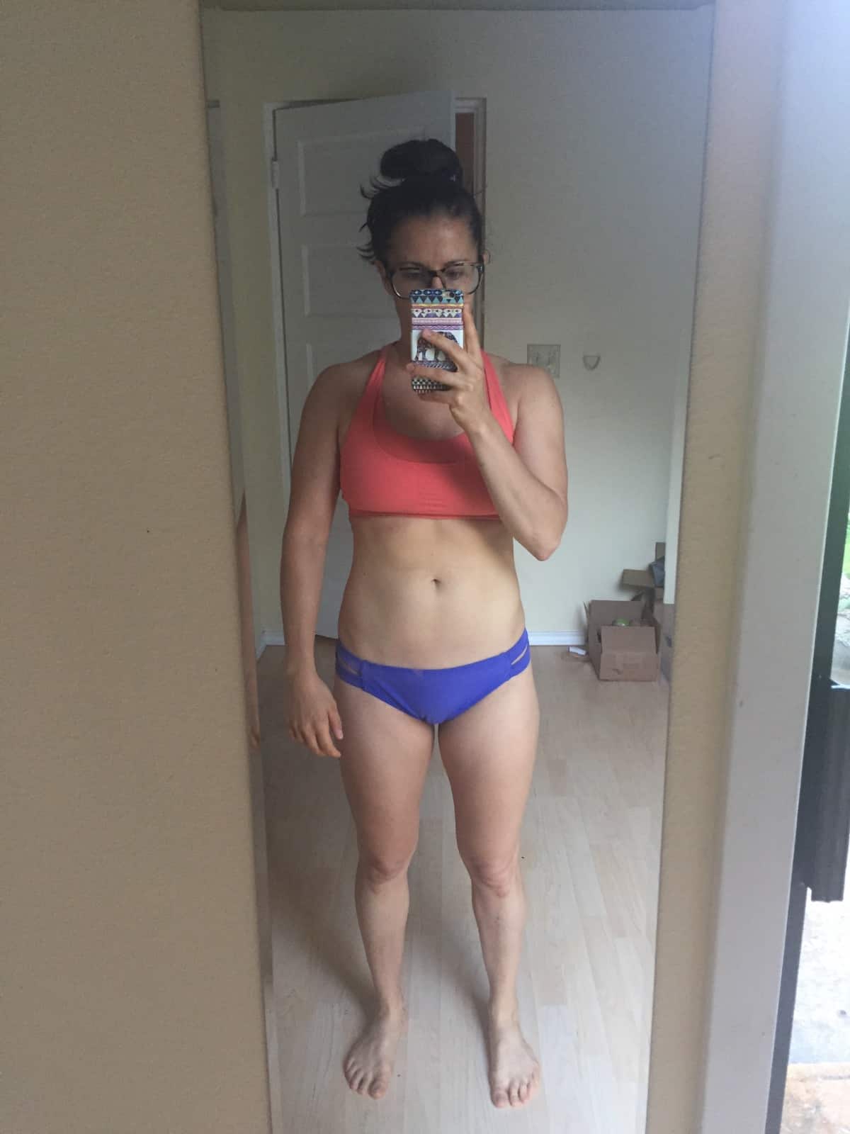 Girl in a bikini bottom and a sports bra taking a selfie in front of a mirror.