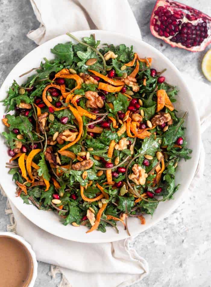 A overhead view of a large white bowl filled with a vegan kale salad with butternut squash and pomegranates. Next to it is a small bowl of tahini dressing, a quarter of a pomegranate, and half a lemon.