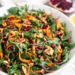 Large white bowl filled with a paleo kale salad with butternut squash and pomegranates. It also has walnuts and pumpkin seeds in it. Behind the bowl is a quarter of a pomegranate, half a lemon, and a small bowl of dressing further behind them.