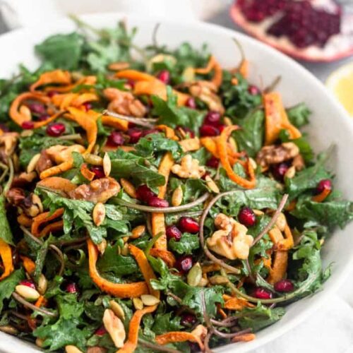 Large white bowl filled with a paleo kale salad with butternut squash and pomegranates. It also has walnuts and pumpkin seeds in it. Behind the bowl is a quarter of a pomegranate, half a lemon, and a small bowl of dressing further behind them.