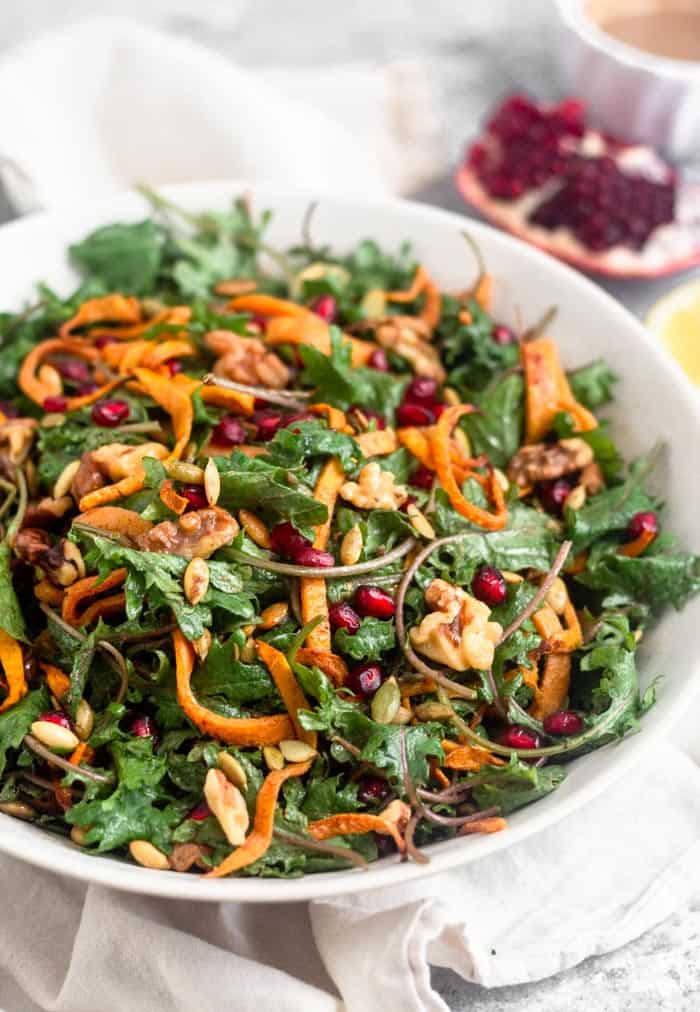Large white bowl filled with a paleo kale salad with butternut squash and pomegranates. It also has walnuts and pumpkin seeds in it. Behind the bowl is a quarter of a pomegranate, half a lemon, and a small bowl of dressing further behind them.