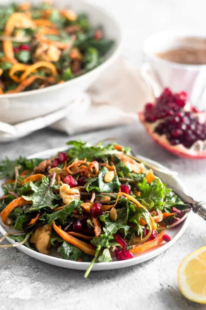 Small plate filled with a Whole30 kale salad with butternut squash and pomegranates with a fork hanging off of it. Behind it is a quarter of a pomegranate, a small bowl of dressing, and a large bowl of more salad. I front of the small plate is half a lemon.
