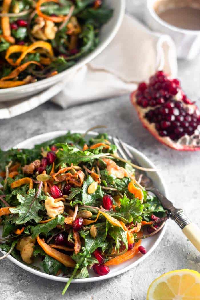 Small white plate filled with kale salad with butternut squash with a fork hanging off of it. Behind it is a quarter of a pomegranate, a small bowl of dressing, and a large bowl of more salad.