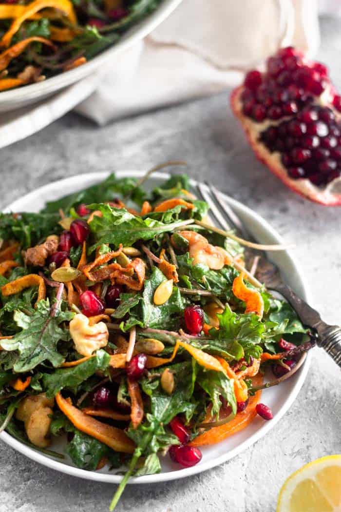 Small plate filled with a vegan kale salad with butternut squash and pomegranates. A fork is hanging off the side and behind it is a large bowl of salad and a quarter of an opened pomegranate.