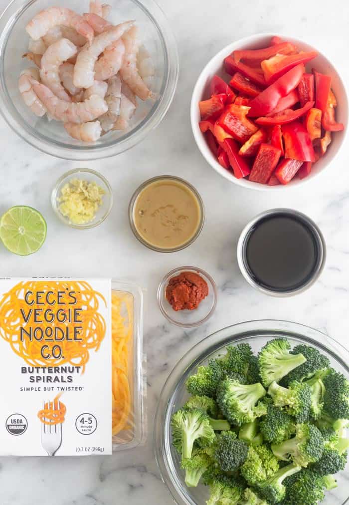 The ingredients for whole30 shrimp & butternut squash noodles with curry peanut sauce on a white counter. There is peeled shrimp, sliced red pepper, coconut aminos, broccoli, curry paste, sunbutter, ginger, garlic, and butternut squash noodles.