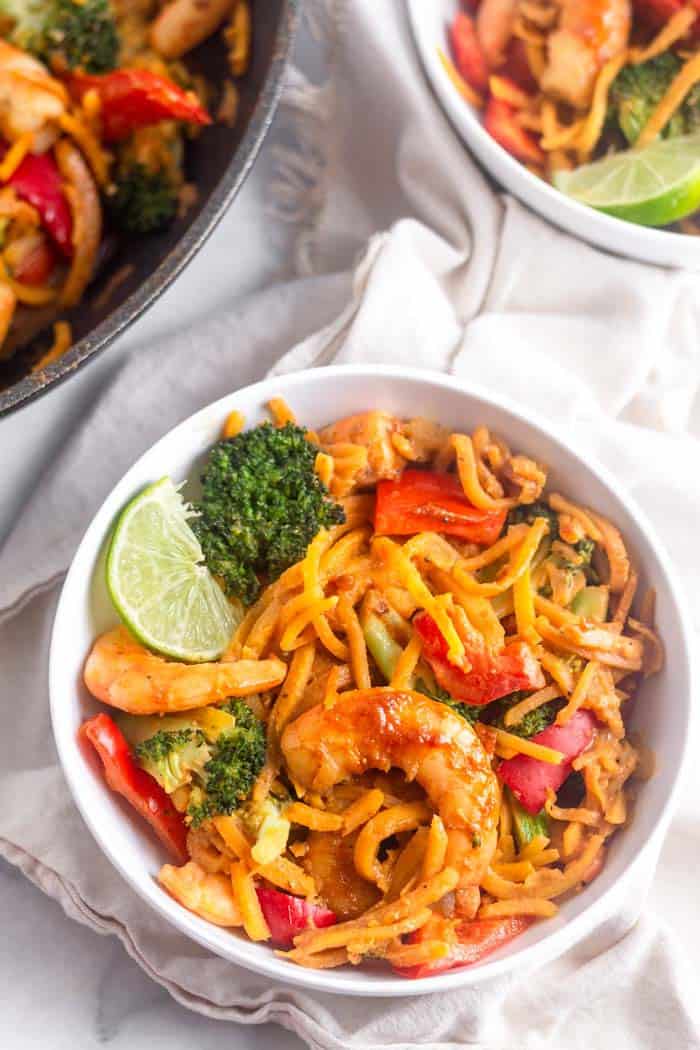 Overhead shot of a bowl filled with Whole30 Shrimp & Butternut Squash Noodles with Curry Peanut Sauce garnished with a lime wedge. It is sitting on a tan linen and next to it is another full bowl and a the pan of noodles.